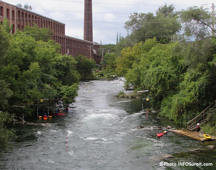 Photo : rivière naturelle un peu mouvementée ; forêt verte touffue autour, longée par une usine en briques rouges et à grande cheminée ; des amateur.trices de Kayak sont visibles.